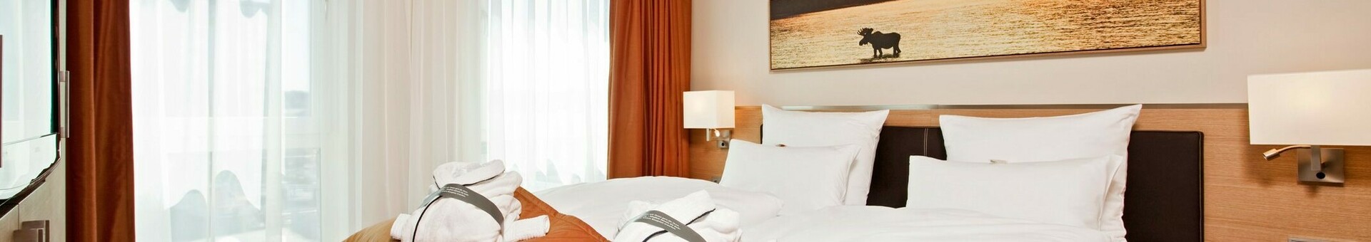 Interior view of the Suite in the ATLANTIC Hotel Kiel with double bed and luxourious furniture Modern hotel room with double bed, white sheets, orange blanket and bathrobes, light-colored curtains.
