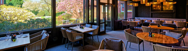 Wintergarten im ATLANTIC Hotel Landgut Horn Gemütliches Restaurant mit Holztischen und -decken, großen Fenstern mit Gartenblick und stilvollen Lampen.