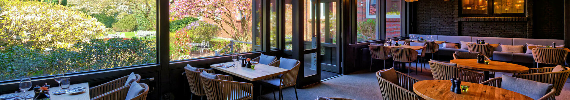 Wintergarten im ATLANTIC Hotel Landgut Horn Gemütliches Restaurant mit Holztischen und -decken, großen Fenstern mit Gartenblick und stilvollen Lampen.