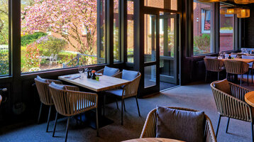 Wintergarten im ATLANTIC Hotel Landgut Horn Gemütliches Restaurant mit Holztischen und -decken, großen Fenstern mit Gartenblick und stilvollen Lampen.