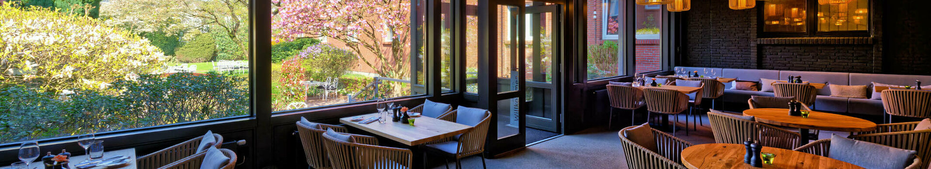 Gemütliches Restaurant mit Holztischen und -decken, großen Fenstern mit Gartenblick und stilvollen Lampen.