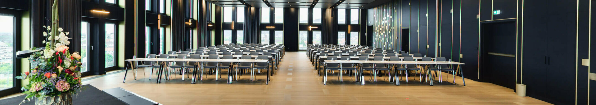 Modern conference room with large windows, rows of tables and chairs, flower arrangement on stage.