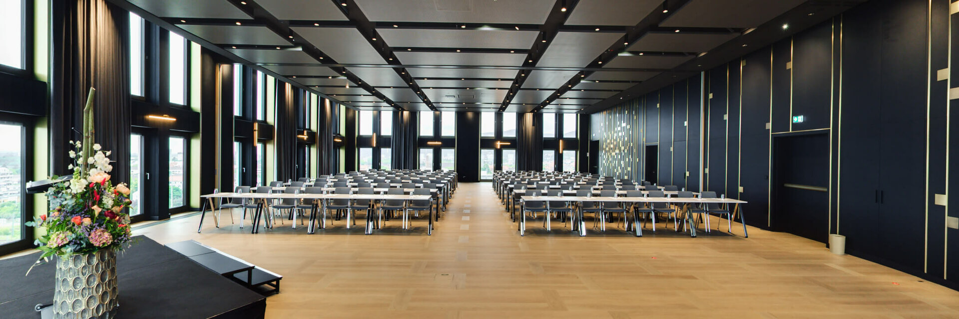 Modern conference room with large windows, rows of tables and chairs, flower arrangement on stage.