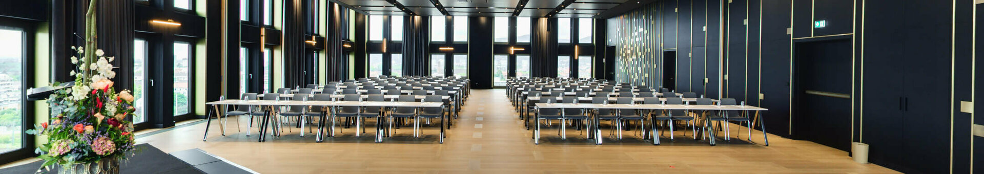 ATLANTIC Hotel Heidelberg Ballsaal parlamentarisch Moderner Konferenzraum mit großen Fenstern, Reihen von Tischen und Stühlen, Blumenarrangement auf Bühne.