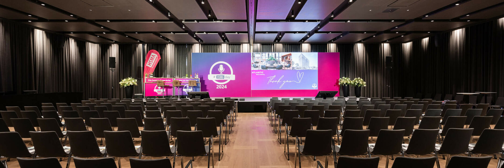 Large conference room in the ATLANTIC Hotel Heidelberg with rows of chairs and illuminated podium.