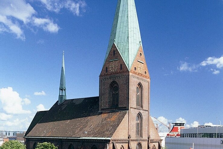 Große Backsteinkirche mit grünem Turmdach, umgeben von modernem Einkaufszentrum und belebtem Platz bei blauem Himmel.