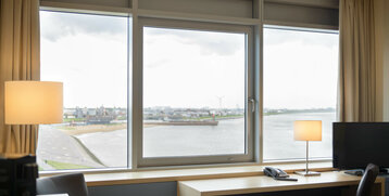 Blick aus einem Hotelzimmerfenster auf die Weser und Bremerhaven, mit Schreibtisch, Lampe und Telefon im Vordergrund.