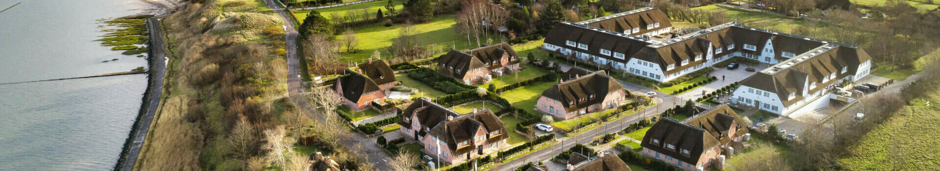 Severins Resort & Spa Sylt Aussenaufnahme Aerial view of a hotel with thatched roof houses, surrounded by green meadows and close to the coast.