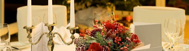 Festlich gedeckter Tisch mit Kerzenleuchter, roten Blumen und Weihnachtsbaum im Hintergrund.