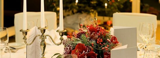 Festlich gedeckter Tisch mit Kerzenleuchter, roten Blumen und Weihnachtsbaum im Hintergrund.