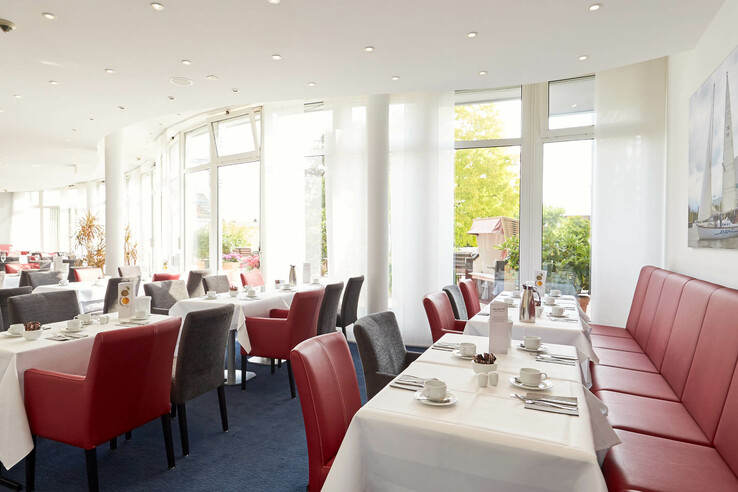 Bright hotel restaurant with red and gray chairs, white-clothed tables and large windows.