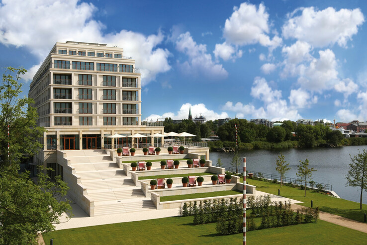 Modernes Hotel am Fluss mit Terrassen, Liegestühlen und Sonnenschirmen, umgeben von gepflegtem Rasen und Bäumen.