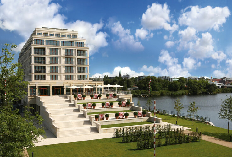 Modernes Hotel am Fluss mit Terrassen, Liegestühlen und Sonnenschirmen, umgeben von gepflegtem Rasen und Bäumen.