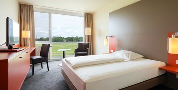 Bright hotel room with double bed, desk, armchair and large window with a view of the racecourse.