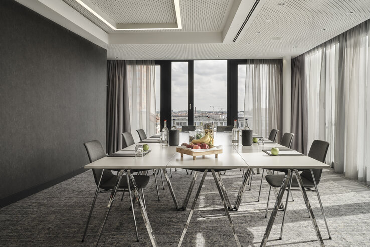 Modern conference room in the ATLANTIC Hotel Münster with large table, chairs, water bottles and fruit, bright window front.