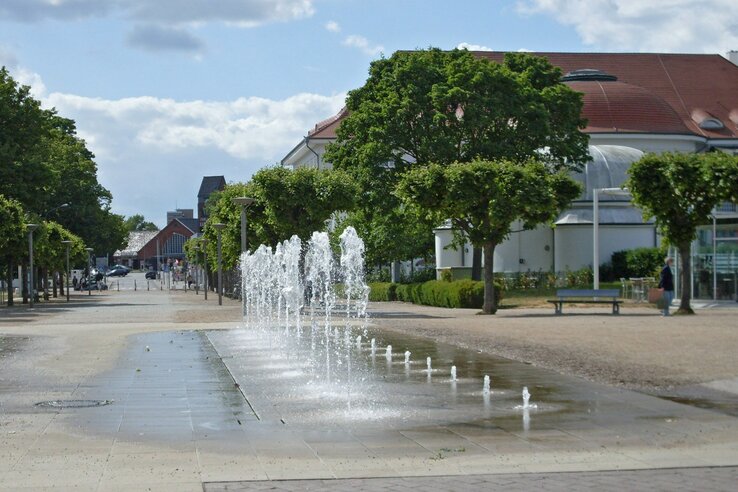 Springbrunnen auf Promenade vor dem ATLANTIC Grand Hotel Travemünde, umgeben von Bäumen und Gebäuden bei sonnigem Wetter.