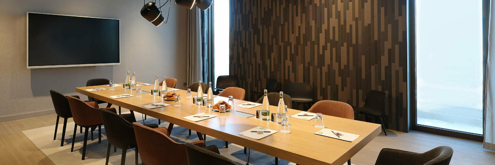Moderner Konferenzraum mit großem Holztisch, Stühlen, Flachbildschirm und dekorativer Wand im ATLANTIC Hotel Heidelberg.