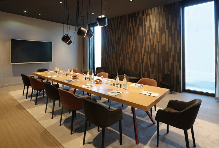 Moderner Konferenzraum mit großem Holztisch, Stühlen, Flachbildschirm und dekorativer Wand im ATLANTIC Hotel Heidelberg.