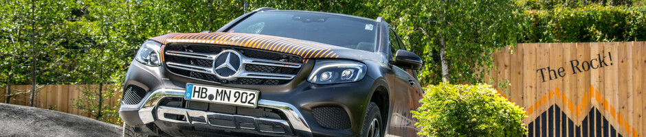 SUV drives over off-road obstacle in front of wooden fence labeled "The Rock!", surrounded by trees and bushes.