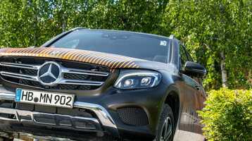 SUV fährt über Offroad-Hindernis vor Holzzaun mit Aufschrift "The Rock!", umgeben von Bäumen und Sträuchern.