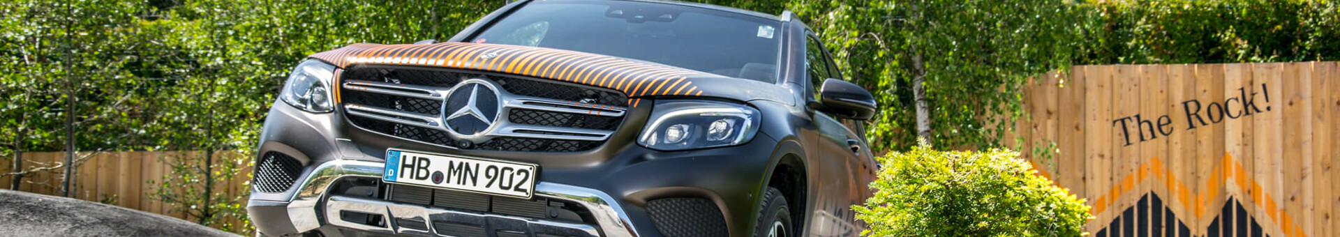 SUV drives over off-road obstacle in front of wooden fence labeled "The Rock!", surrounded by trees and bushes.