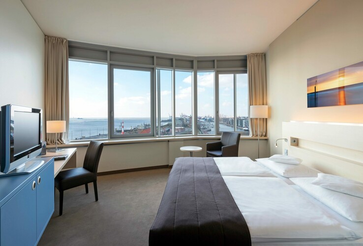 Helles Hotelzimmer mit Doppelbett, Schreibtisch, TV und Panoramafenster mit Blick auf Hafen und Meer in Bremerhaven.