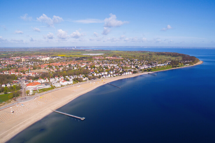 Luftaufnahme des Strandes von Travemünde mit Promenade, Meer und angrenzender Stadt in der Ferne bei klarem Himmel.