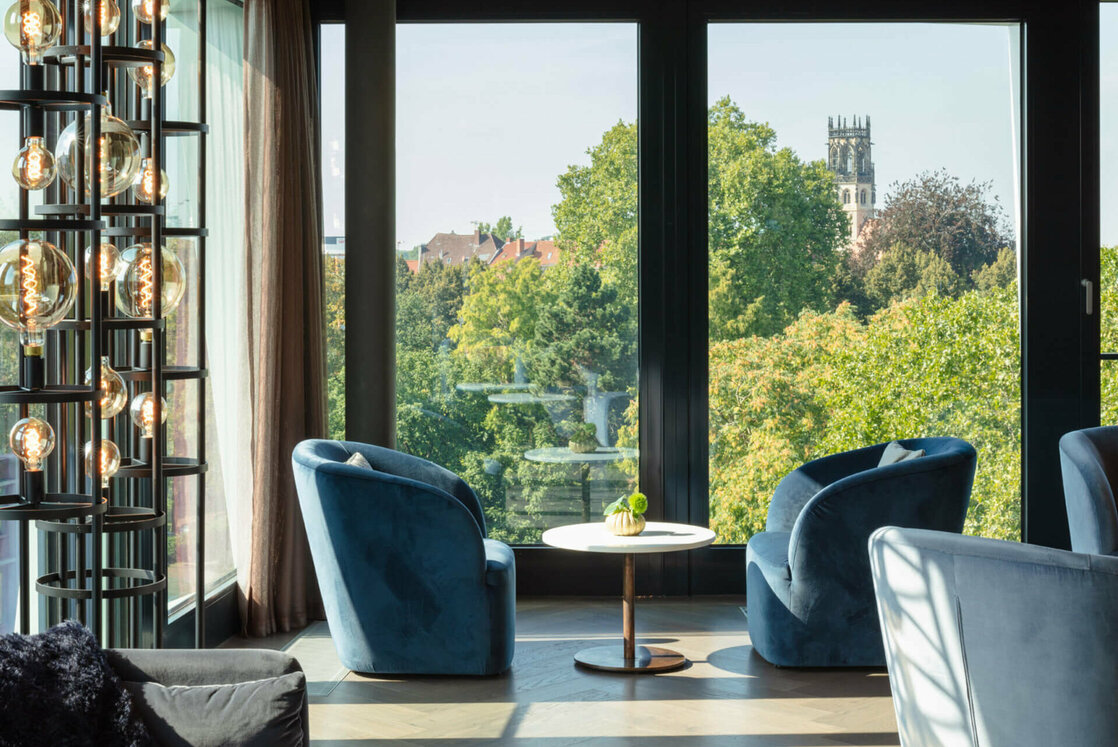 ATLANTIC Skybar Münster | Bar des Jahres 2022 Moderner Lounge-Bereich mit blauen Sesseln, großem Fenster und Blick auf grüne Bäume und Kirchturm.