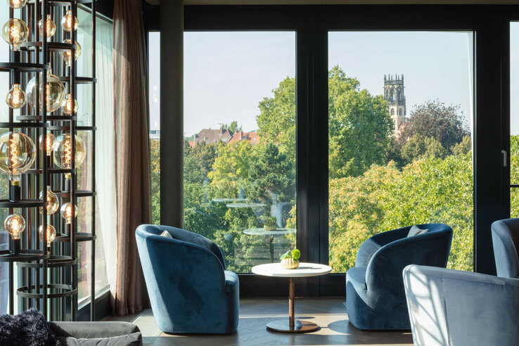 Modern lounge area with blue armchairs, large window and view of green trees and church tower.