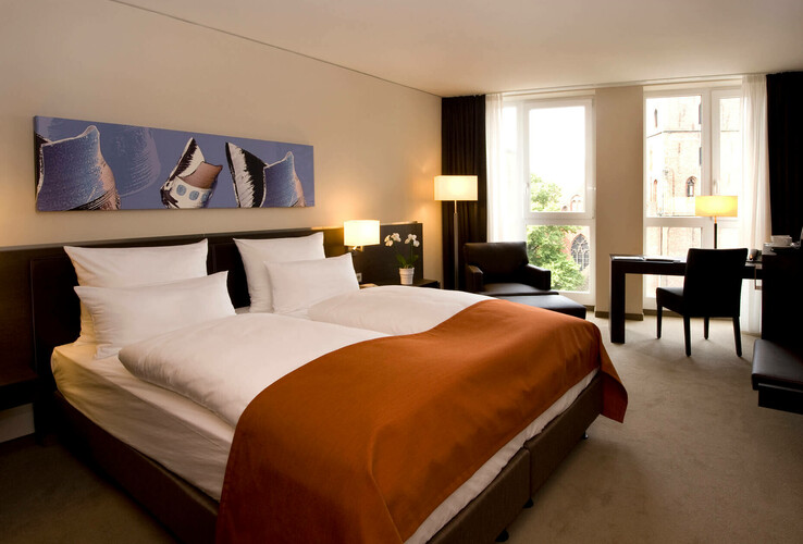 Modernes Hotelzimmer mit Doppelbett, orangefarbener Decke, Schreibtisch, Sesseln und großem Fenster mit Blick auf eine Kirche.