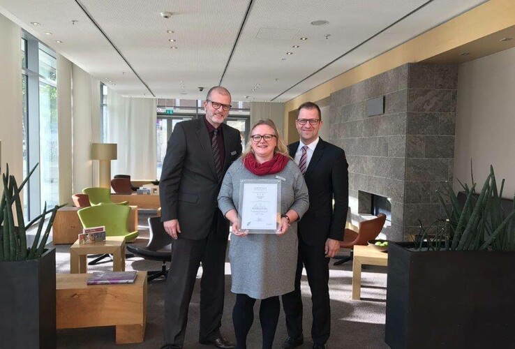 Three people are standing in a modern hotel lobby, one woman is holding an award.