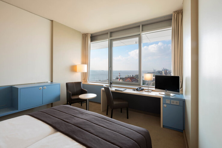 ATLANTIC Hotel SAIL City – Comfort Plus Room Bright hotel room with sea view, desk, chair, bed and blue accents at the ATLANTIC Hotel Sail City, Bremerhaven.