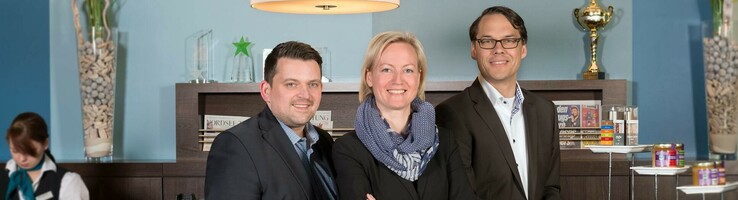 Three hotel employees in suits smile at the reception desk of the ATLANTIC Hotel Sail City, Bremerhaven.