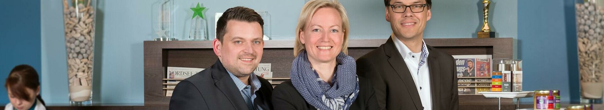 Three hotel employees in suits smile at the reception desk of the ATLANTIC Hotel Sail City, Bremerhaven.