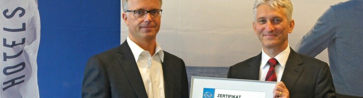 Geschäftsführer der ATLANTIC Hotels mit Zertifizierung in der Hand Zwei Männer in Anzügen präsentieren ein Zertifikat vor einem Banner der Atlantic Hotels.