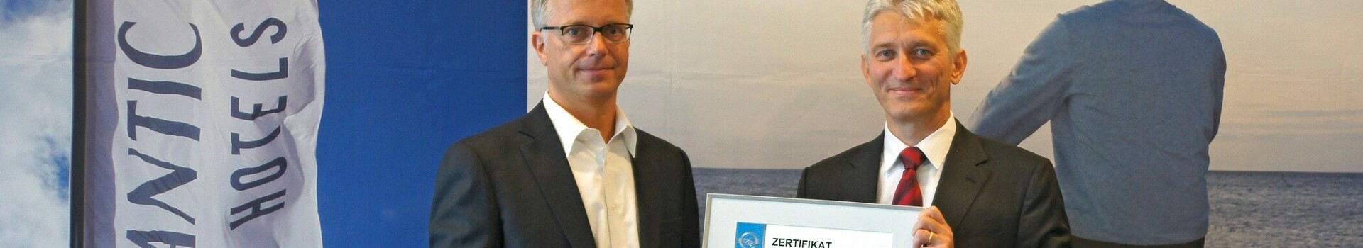 Zwei Männer in Anzügen präsentieren ein Zertifikat vor einem Banner der Atlantic Hotels.