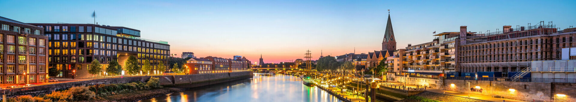 Riverbank at sunset, illuminated buildings and promenade reflected in the water.