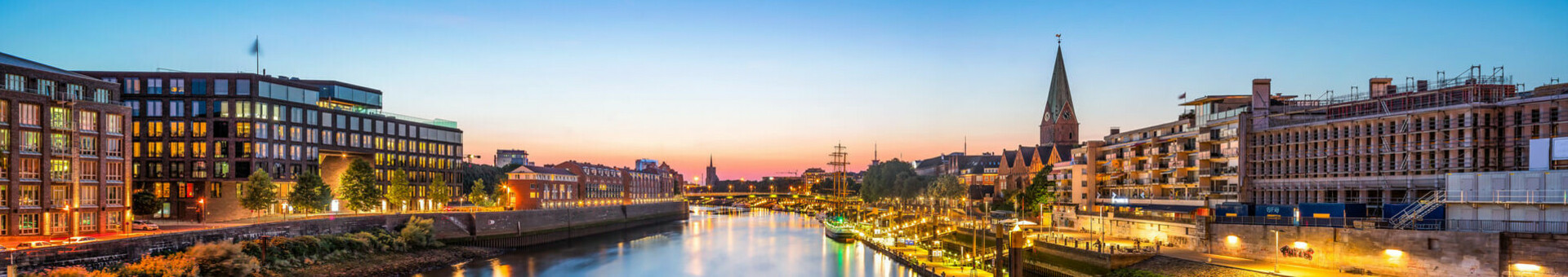 Flussufer bei Sonnenuntergang, beleuchtete Gebäude und Promenade spiegeln sich im Wasser.