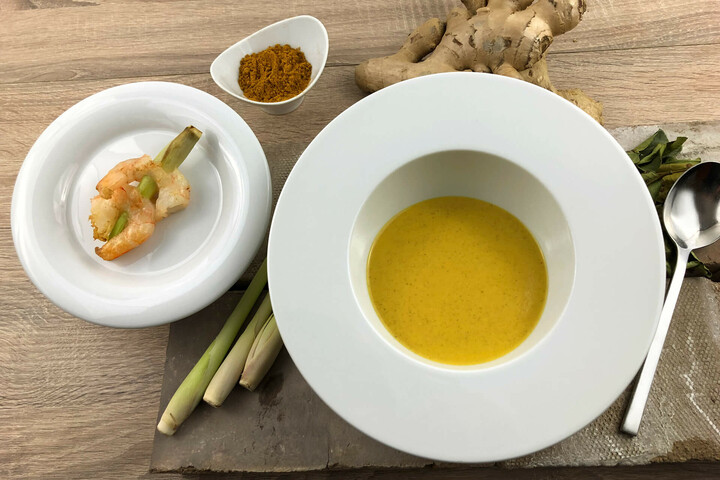 Yellow soup in a white plate, next to it a prawn skewer, curry powder, ginger and lemongrass on a wooden table.