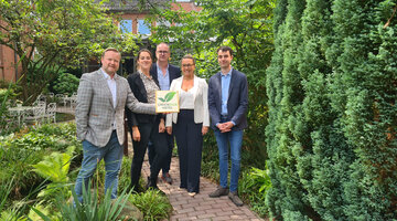 Pressemitteilung | ATLANTIC Hotels Gruppe GreenSign zertifiziert Fünf Personen stehen in einem grünen Garten und halten ein Schild mit der Aufschrift "Greensign Hotel".