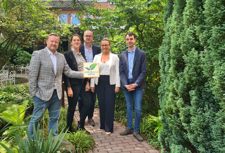 Pressemitteilung | ATLANTIC Hotels Gruppe GreenSign zertifiziert Fünf Personen stehen in einem grünen Garten und halten ein Schild mit der Aufschrift "Greensign Hotel".