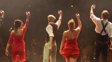 Eine Gruppe von acht Künstlern hebt auf einer Bühne feierlich Gläser vor einem applaudierenden Publikum.