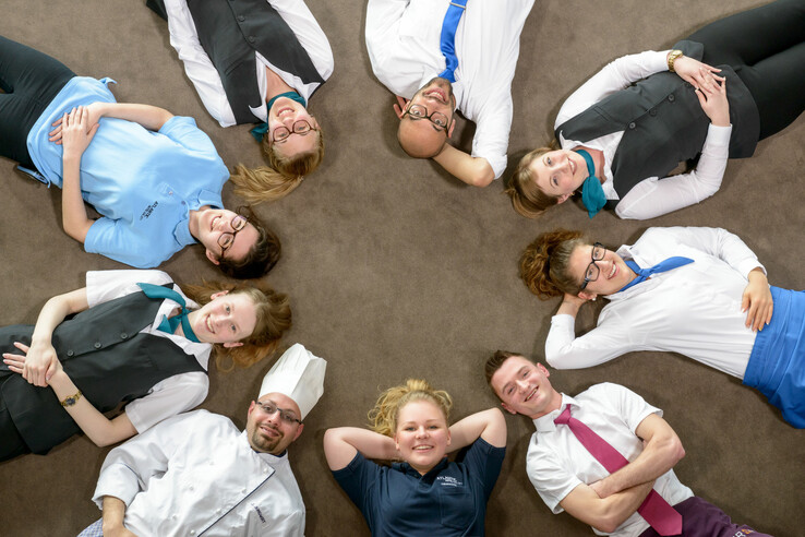 Hotel employees lie smiling in a circle on the floor, wearing different uniforms.