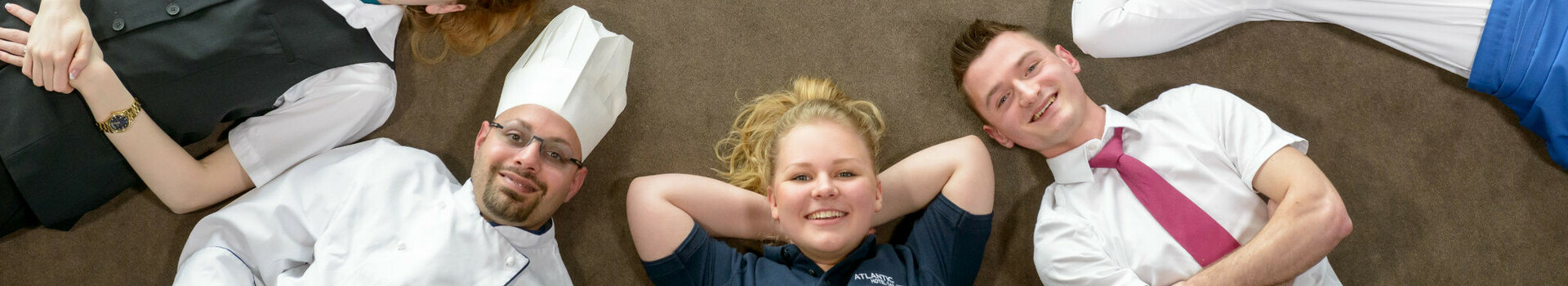 Hotelmitarbeiter liegen lächelnd im Kreis auf dem Boden, tragen verschiedene Uniformen.