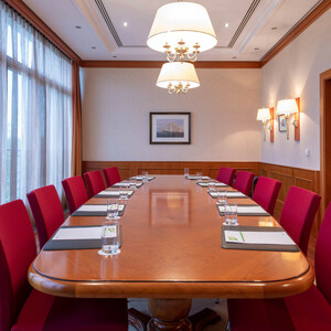 Tagungsraum Münster im ATLANTIC Hotel Wilhelmshaven Eleganter Konferenzraum mit großem Holztisch, roten Stühlen, Notizblöcken und Wassergläsern, beleuchtet durch große Fenster.