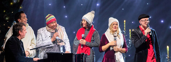 A group of people sing on a stage with festive lighting and decorations.