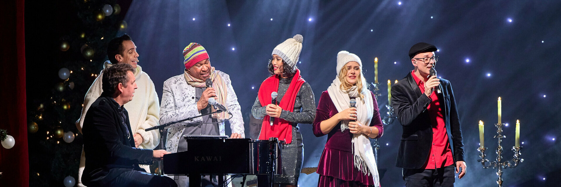 A group of people sing on a stage with festive lighting and decorations.