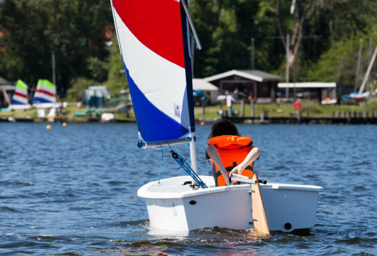 Person sails in a small boat with a red and blue sail on a lake, wearing an orange life jacket.