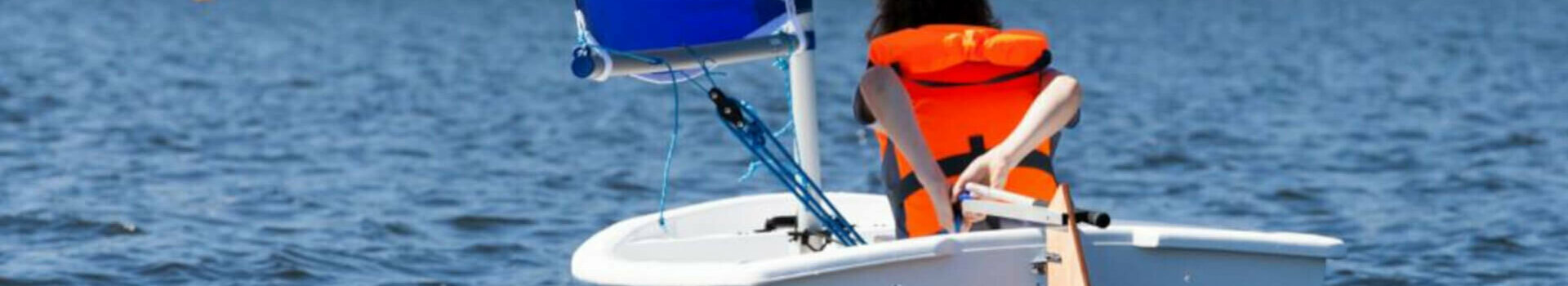 Person sails in a small boat with a red and blue sail on a lake, wearing an orange life jacket.