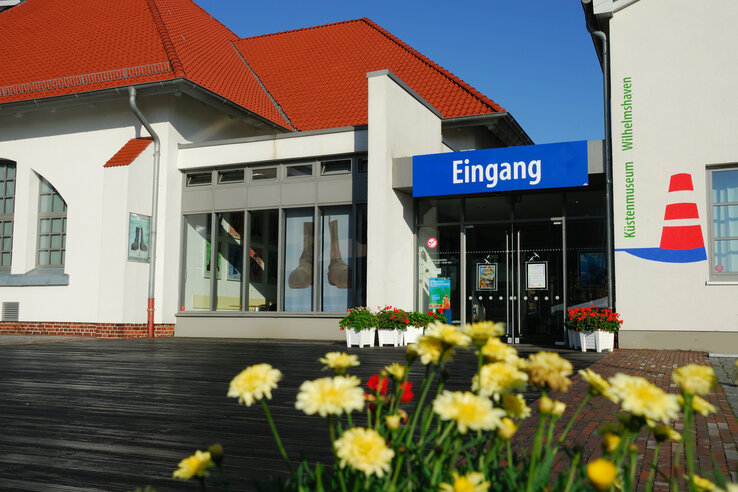 Eingang des Küstenmuseums Wilhelmshaven mit Blumen im Vordergrund und blauem Himmel.